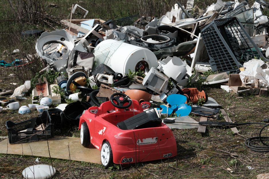 An aerial view showing a large, irregularly shaped pile of assorted rubbish scattered across a patch of land adjacent to a fence and a paved pathway. The trash consists of various materials including 